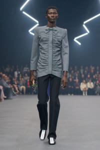 Male model on a runway wearing a light gray ruched long-sleeve top with a bow, black trousers, and white-soled shoes, holding a colorful clutch.
