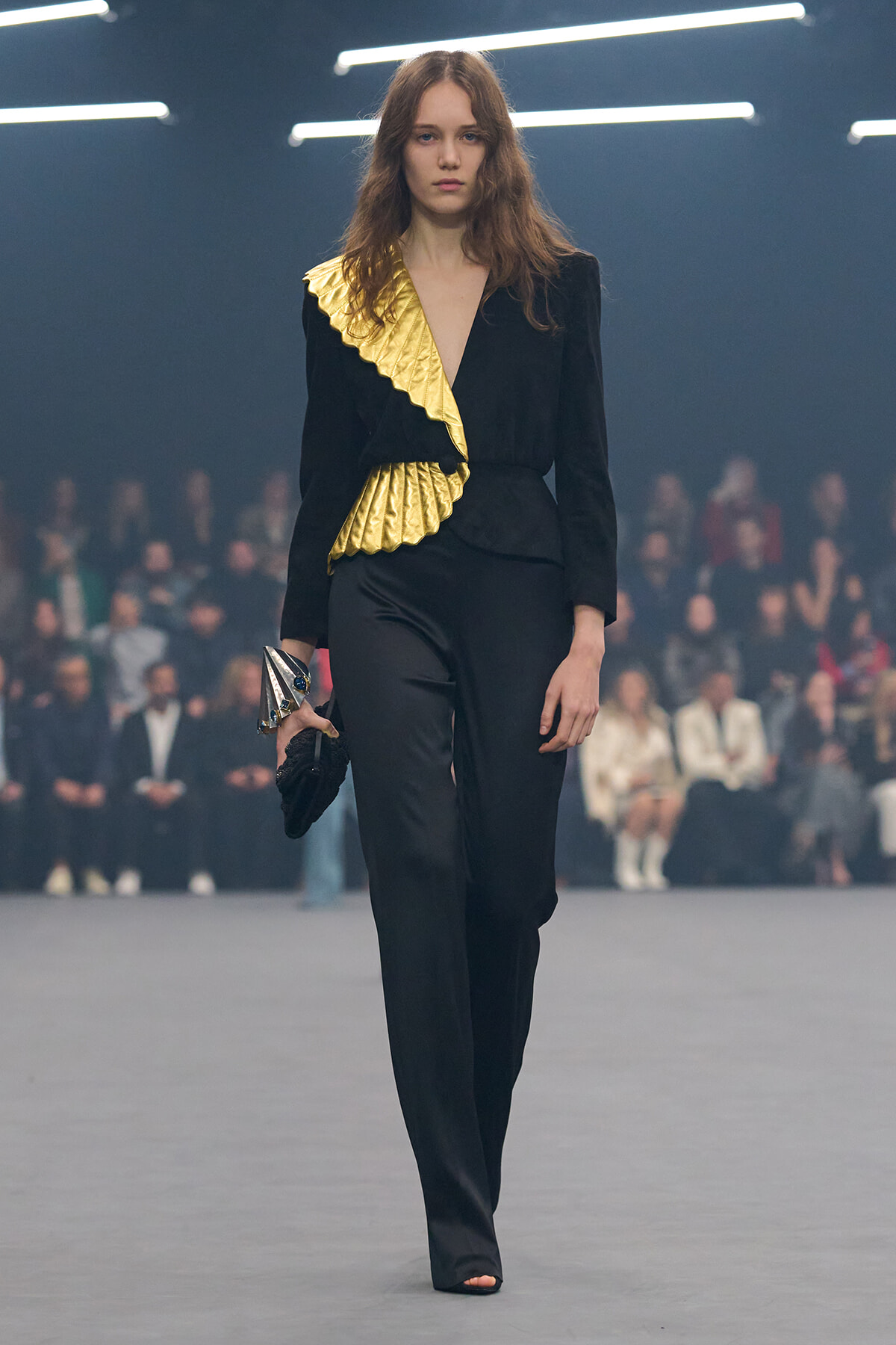 Runway model in a black tailored suit with a gold pleated diagonal sash, holding a metallic clutch as she walks.