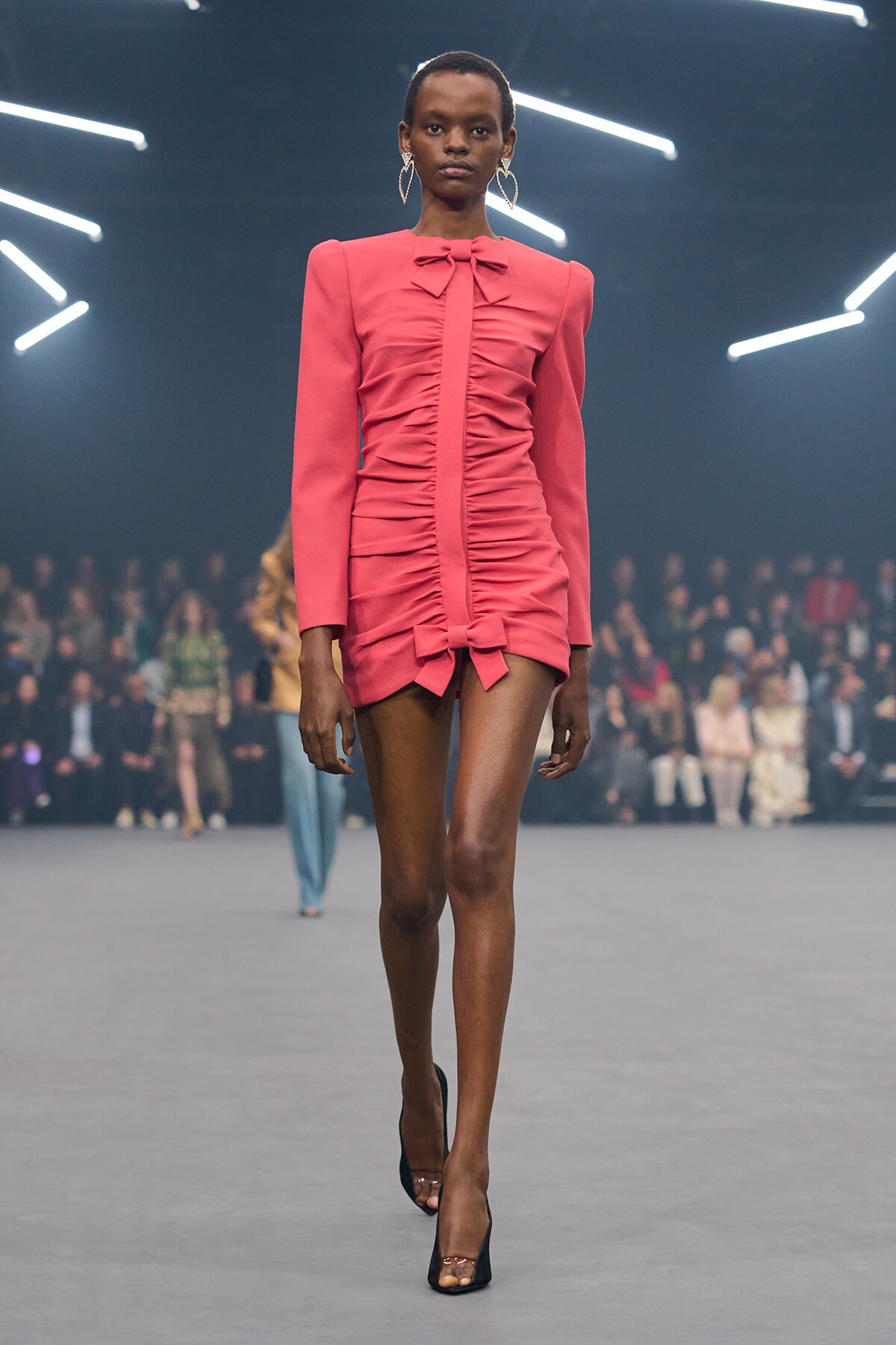 Model wearing a pink ruched mini dress with bow accents walking the runway in black heels.