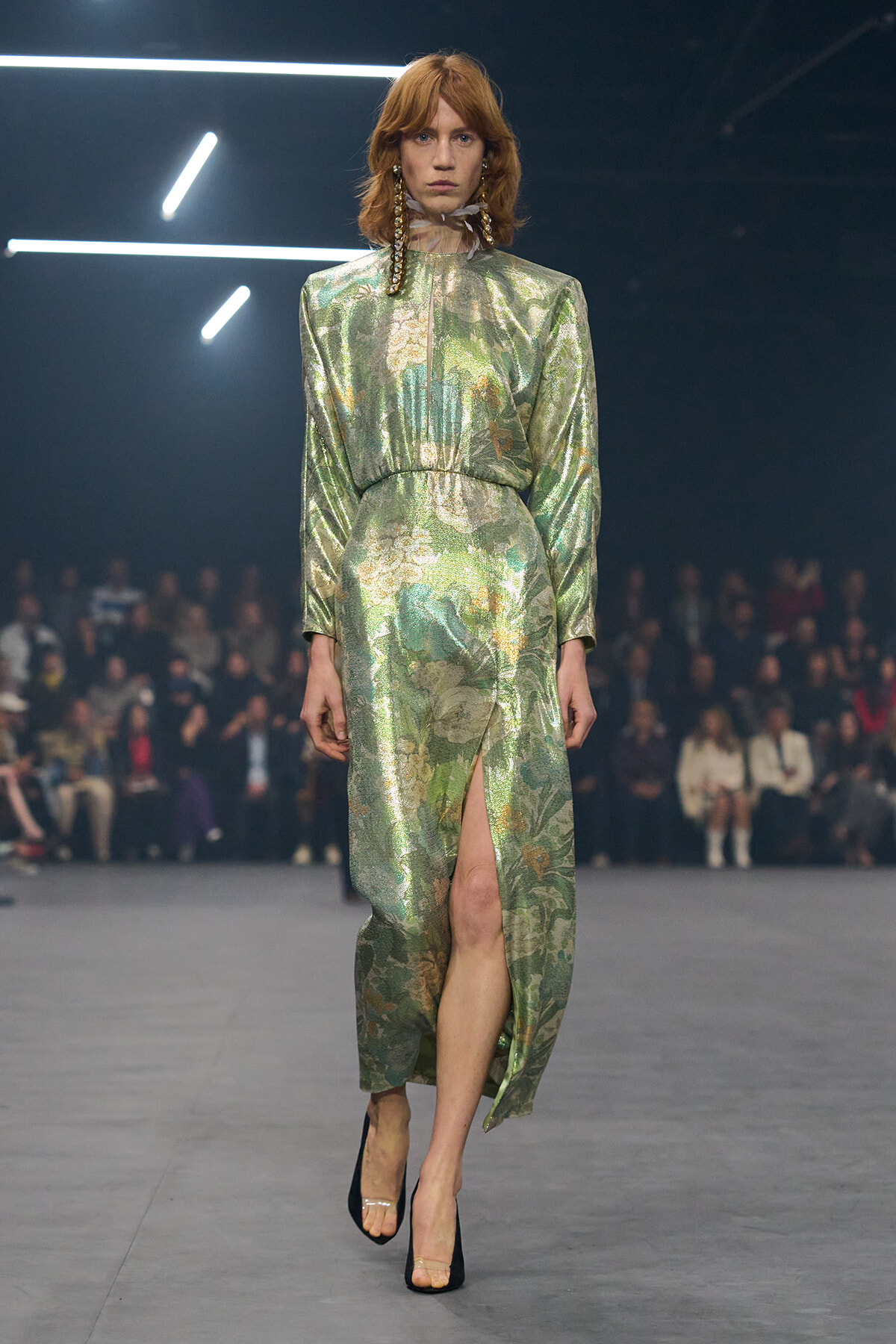 Model in a metallic green floral gown with a high neck and slit, walking a runway with audience in the background.