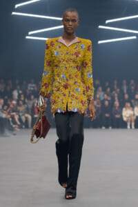 Model walking a fashion runway in a yellow, floral-textured blazer with pink lapels, black trousers, and a burgundy quilted bag on a gold chain.