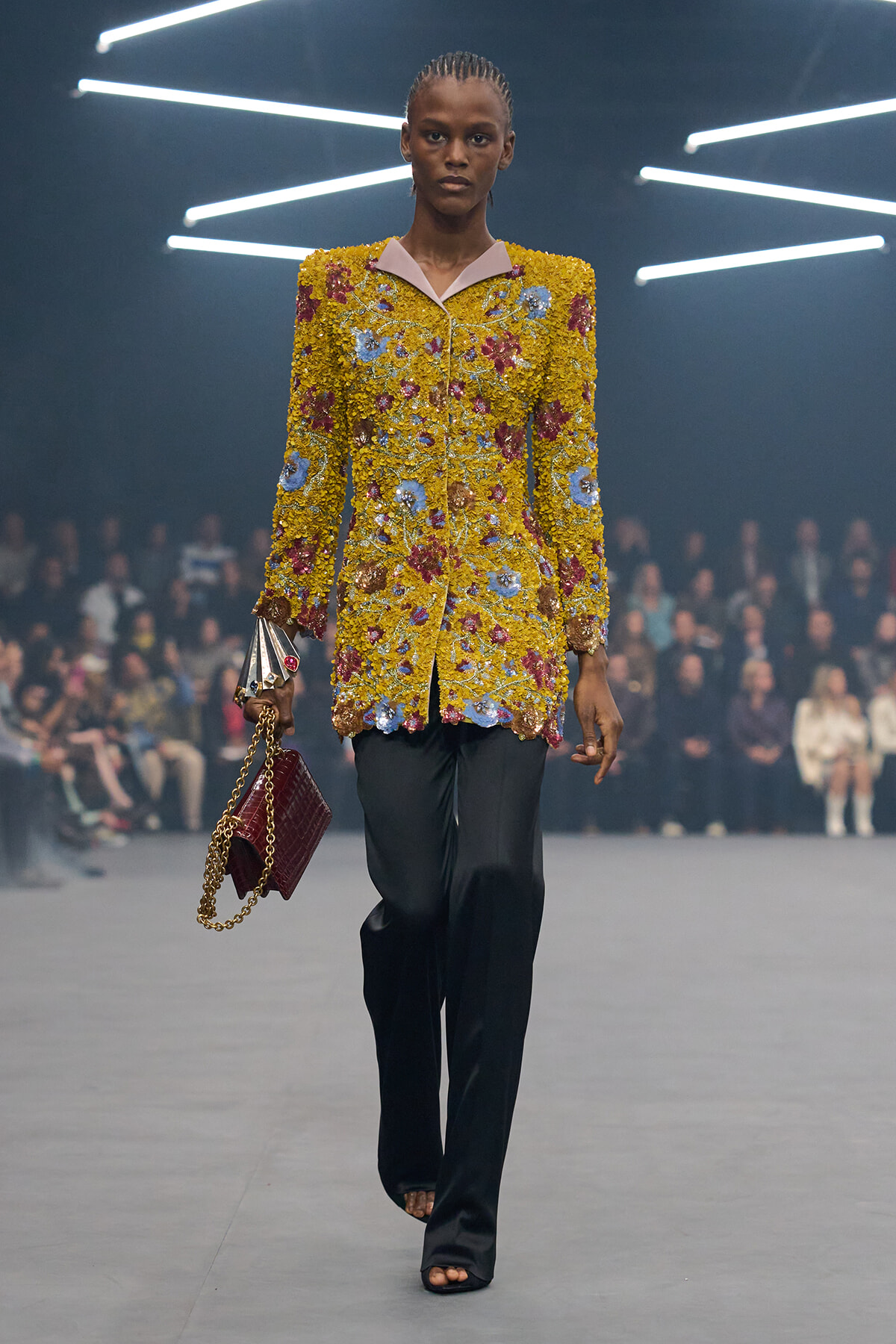 Model walking a fashion runway in a yellow, floral-textured blazer with pink lapels, black trousers, and a burgundy quilted bag on a gold chain.