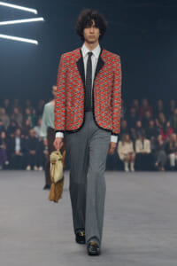 Male runway model wearing a red patterned blazer, white shirt, black tie, gray trousers, and a fringed beige bag in hand on the catwalk.