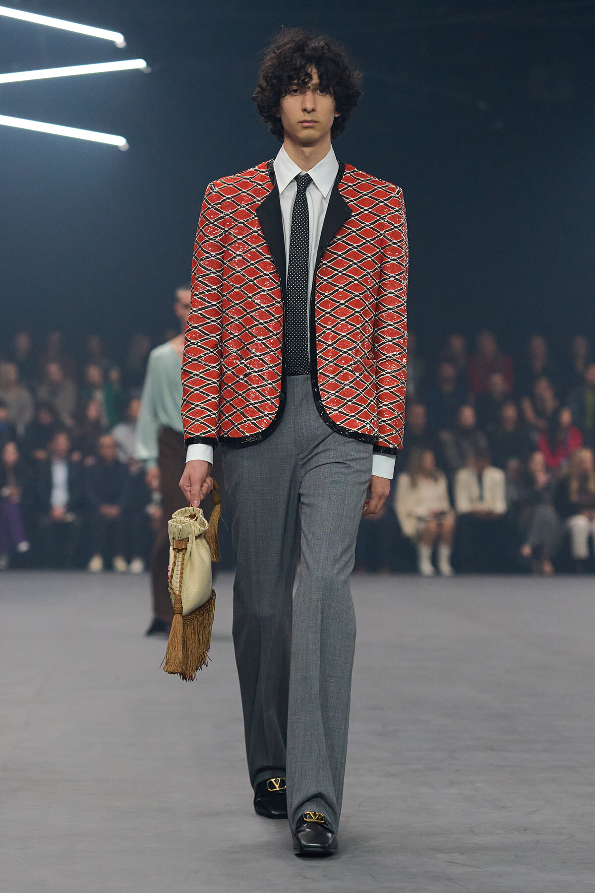 Male runway model wearing a red patterned blazer, white shirt, black tie, gray trousers, and a fringed beige bag in hand on the catwalk.