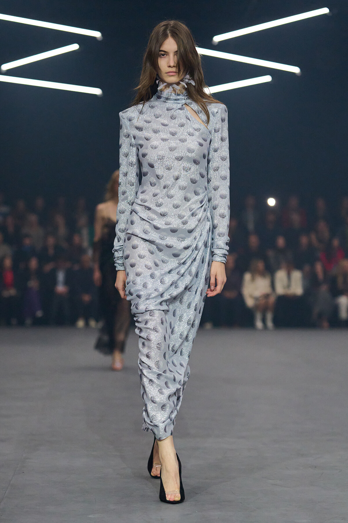 Model walking the runway in a light silver-gray, textured, dotted draped dress with a high neck and cutout detail, black heels.