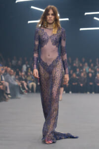 Model walking the runway in a sheer purple lace gown with gold embroidery, in front of a blurred audience.