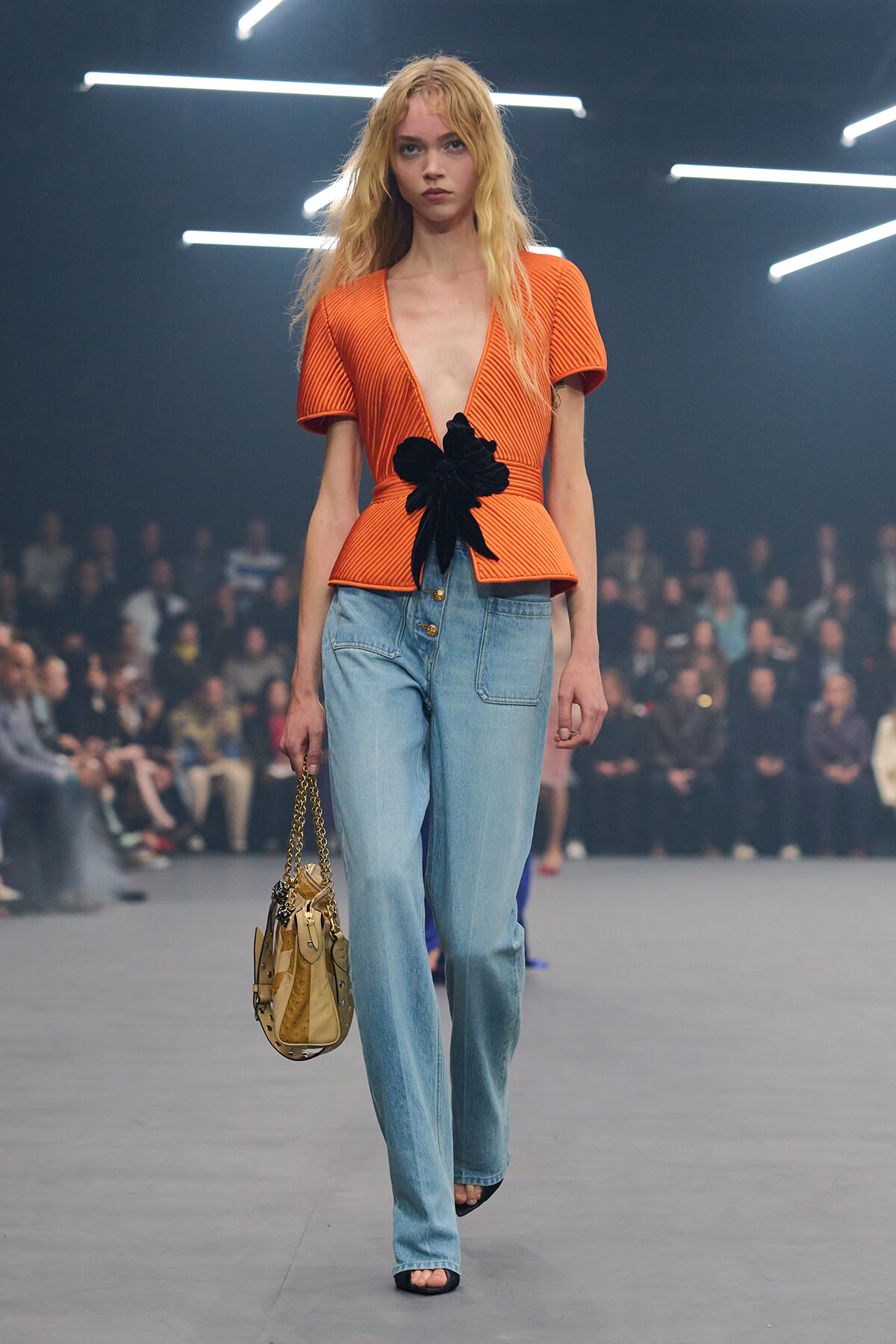 Model walking the runway in an orange ribbed peplum top with a black flower belt, light-blue jeans, and a beige handbag.