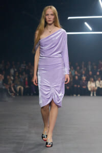 Model walks the runway in a lavender one-shoulder velvet dress with a ruched knee detail, audience in the background.