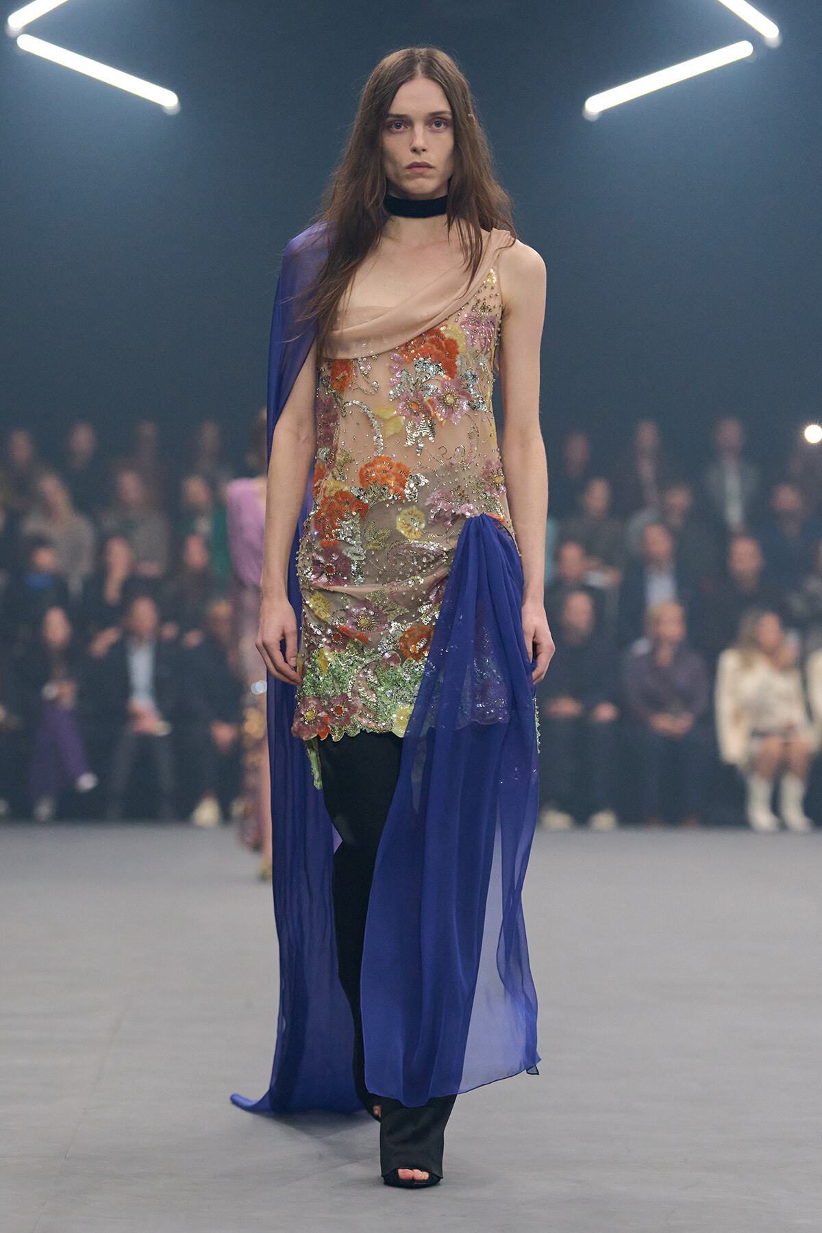 Model walks the runway in a nude-toned, floral-embroidered dress with blue chiffon drape and a black choker.