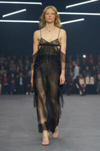 Model walks a fashion runway in a sheer black layered dress with bead trim and off-the-shoulder sleeves.