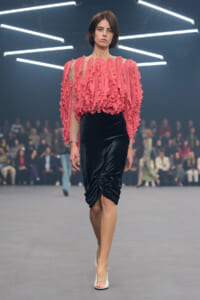 Model walking runway wearing a pink ruffled sheer blouse and black velvet knee-length skirt with white heels.