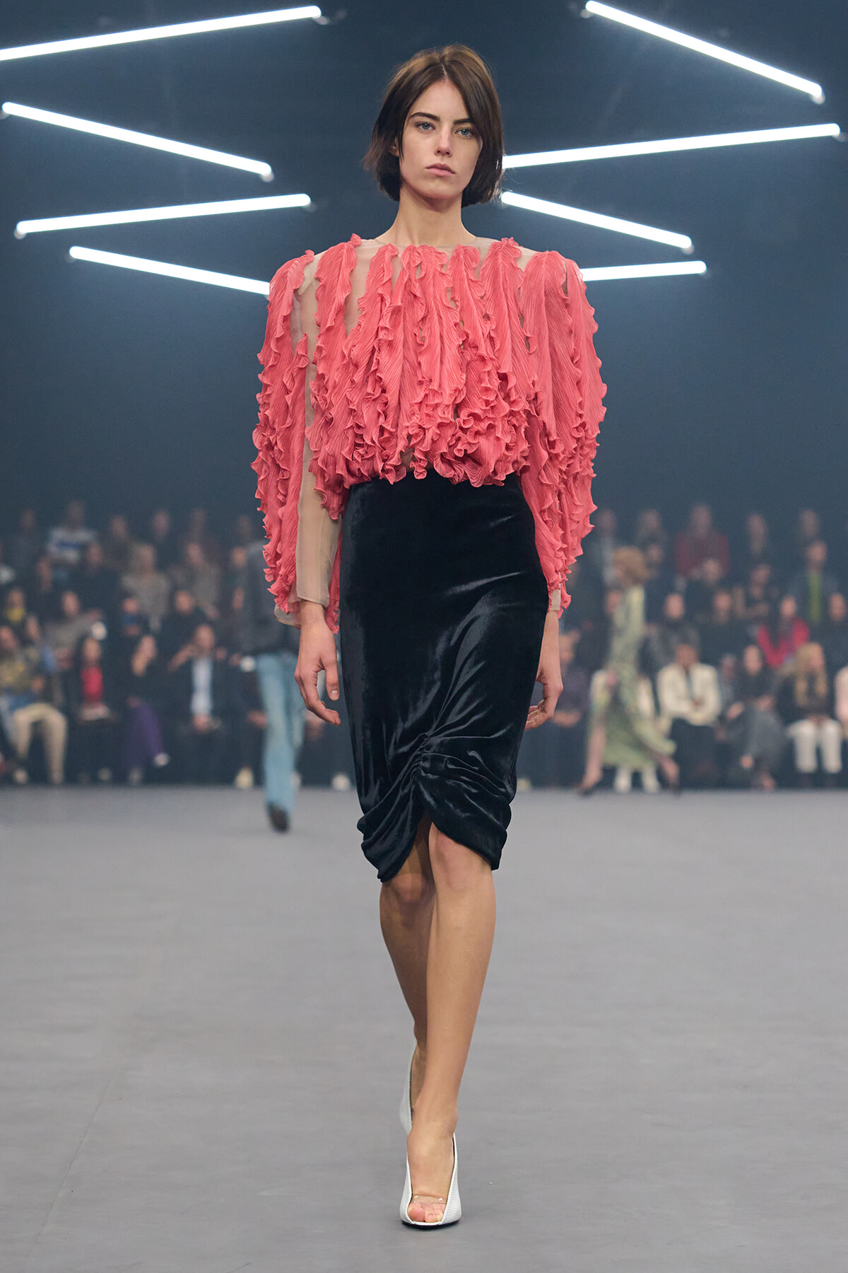 Model walking runway wearing a pink ruffled sheer blouse and black velvet knee-length skirt with white heels.