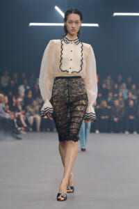 Model on a runway wearing a cream blouse with scalloped black trim and a knee-length black lace pencil skirt.