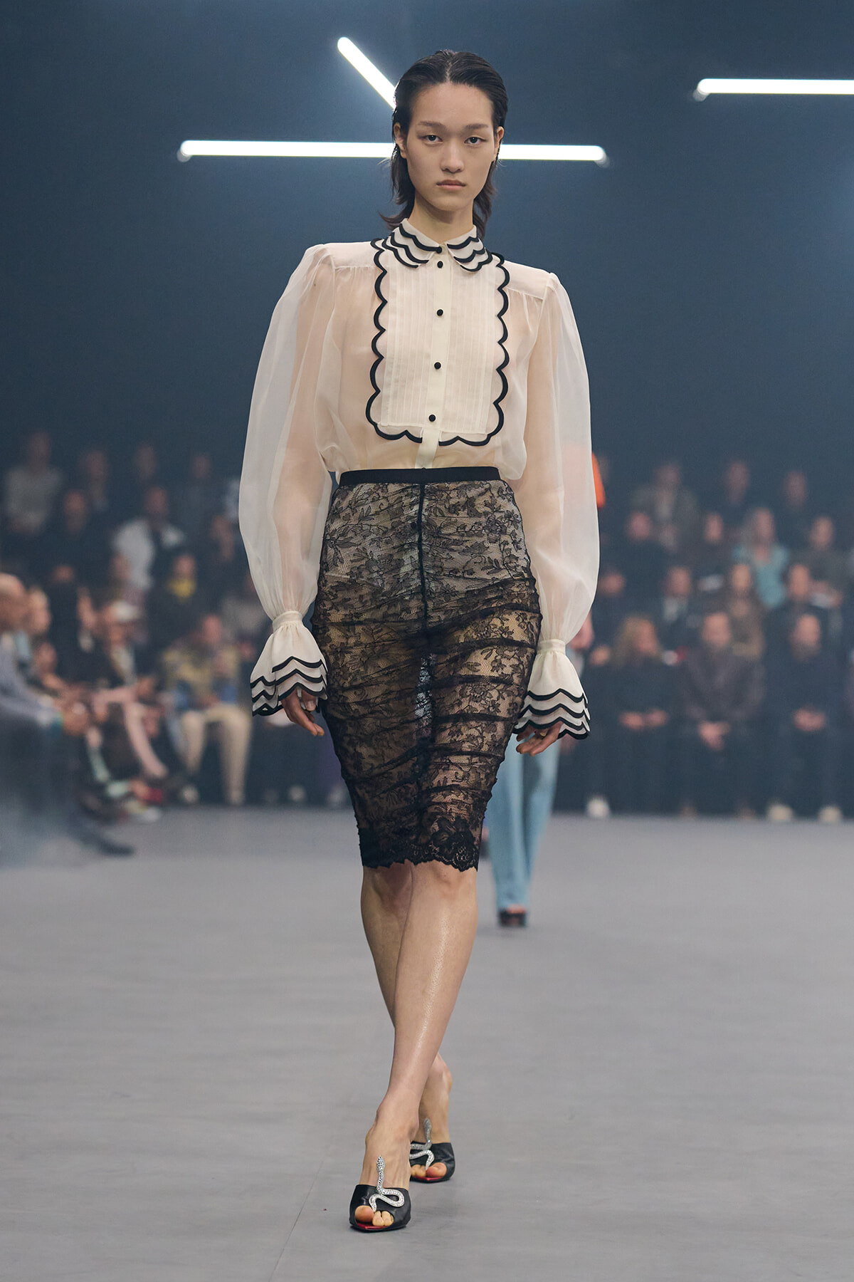 Model on a runway wearing a cream blouse with scalloped black trim and a knee-length black lace pencil skirt.