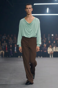 Male model on a runway wearing a mint-green draped blouse and brown wide-leg pants, audience in the background.