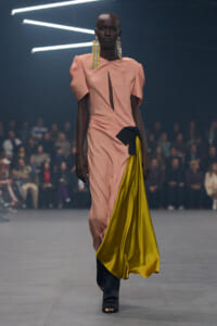 Model walking a runway in a peach dress with puff sleeves and a bold yellow satin drape, plus long gold earrings.