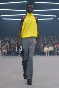 Male model walks a runway in a bright yellow halter top and gray trousers, holding a blue clutch with white embellishment; neon light backdrop and blurred audience.