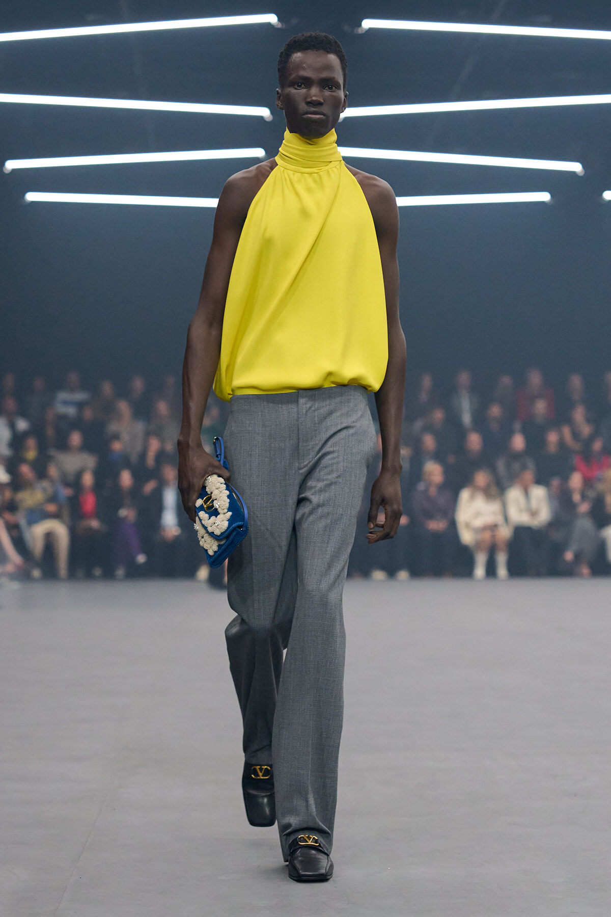 Male model walks a runway in a bright yellow halter top and gray trousers, holding a blue clutch with white embellishment; neon light backdrop and blurred audience.