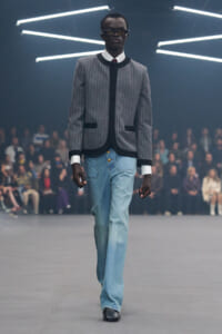 Male model walks a fashion runway in a gray pinstriped jacket with black trim and light blue jeans, audience in the background.