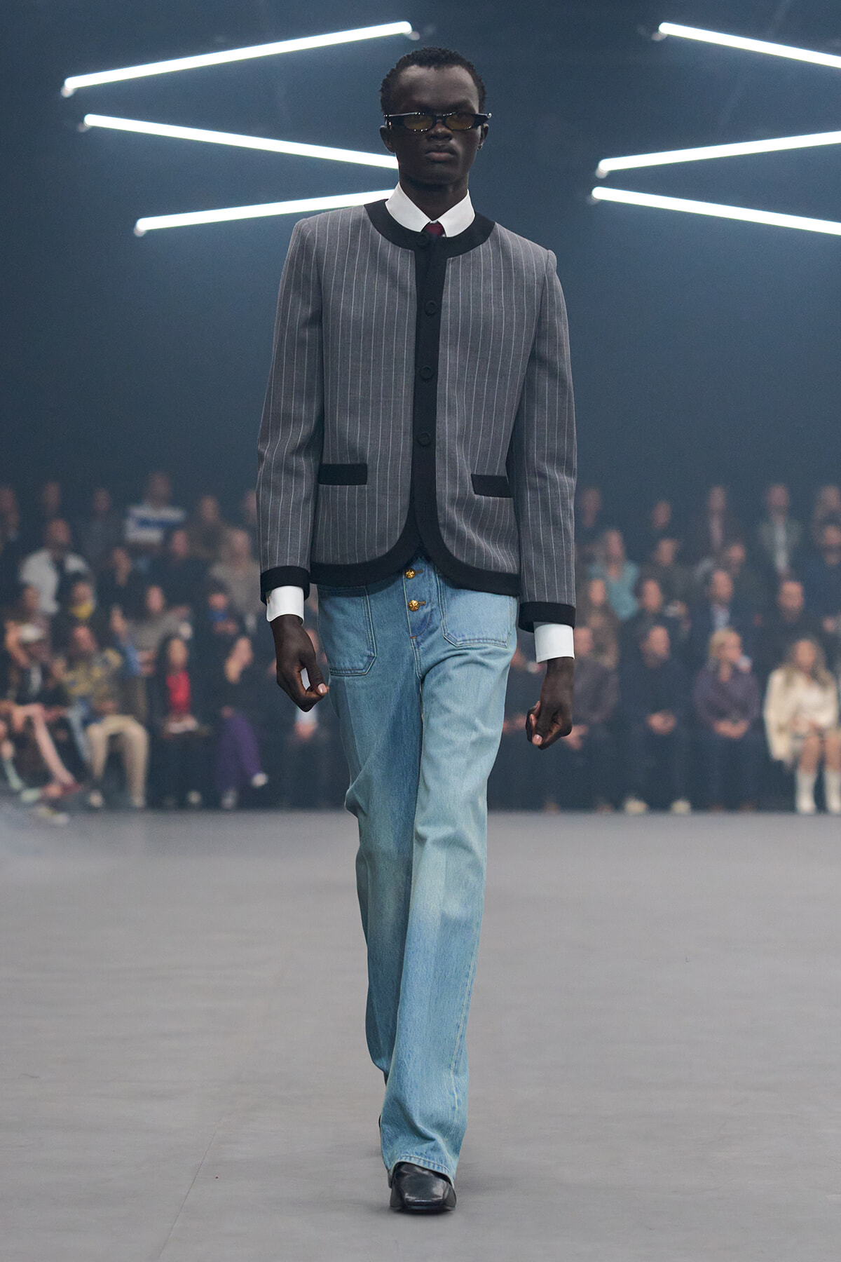 Male model walks a fashion runway in a gray pinstriped jacket with black trim and light blue jeans, audience in the background.