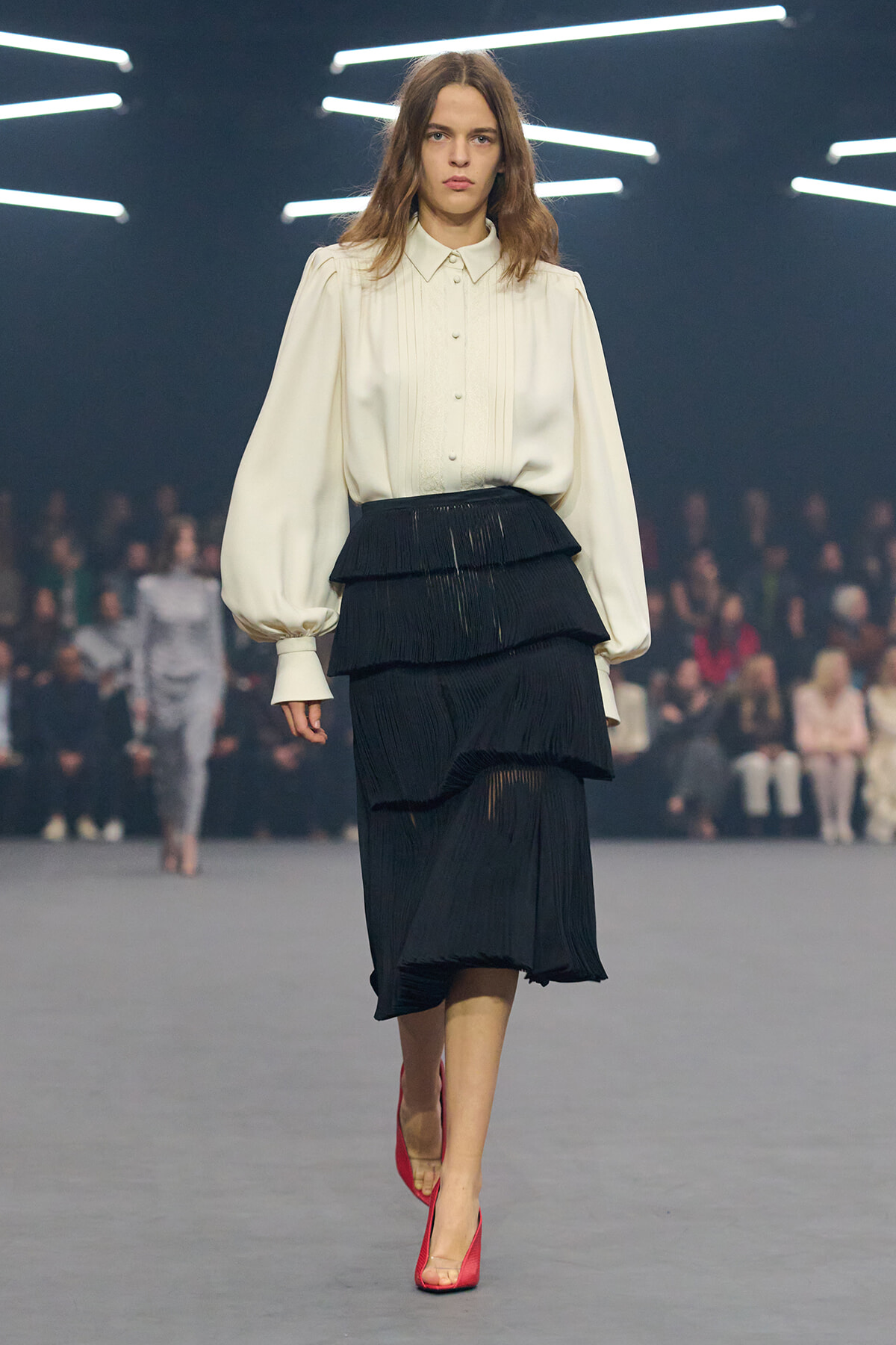 Model walking a fashion runway in a cream blouse with pleated front and layered black tiered skirt, bright red heels.