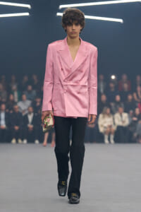 Model walks a fashion runway in a pink satin double-breasted blazer and black trousers, holding a small embroidered clutch.