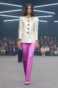 Model on a runway wearing a cream tailored blazer with large shoulders and five black buttons, paired with bright pink satin wide-leg pants.