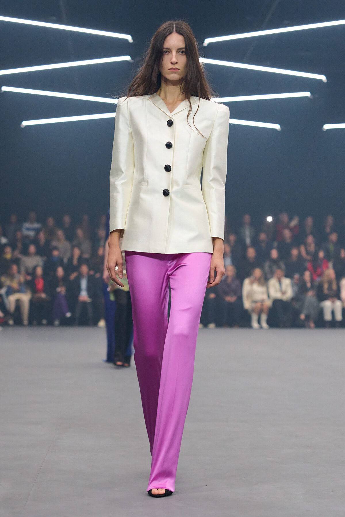 Model on a runway wearing a cream tailored blazer with large shoulders and five black buttons, paired with bright pink satin wide-leg pants.