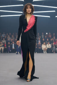 Model walking runway in a black gown with a pink ruffled diagonal panel, thigh-high slit, and gold earrings amid neon-lit backdrop.