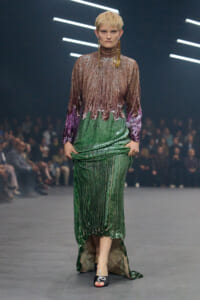 Model with short blond hair walks the runway in a gradient sequin gown: brown top fading to green with purple cuffs, large gold earrings.