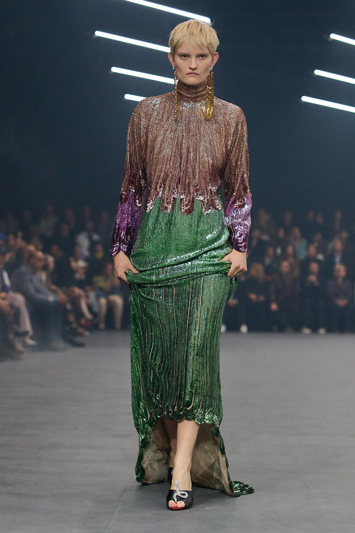 Model with short blond hair walks the runway in a gradient sequin gown: brown top fading to green with purple cuffs, large gold earrings.
