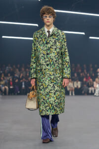 Model walks a fashion show in a knee-length floral coat in greens, yellows, and whites, with a white shirt, tie, purple wide-leg pants, brown shoes, sunglasses, and a beige handbag.