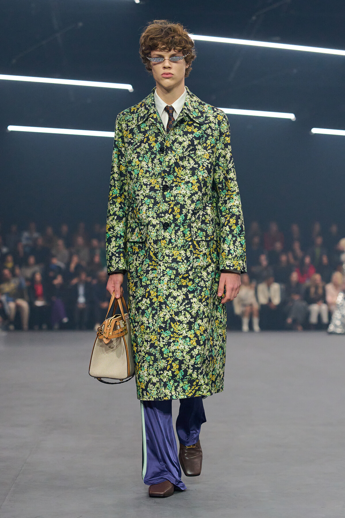 Model walks a fashion show in a knee-length floral coat in greens, yellows, and whites, with a white shirt, tie, purple wide-leg pants, brown shoes, sunglasses, and a beige handbag.
