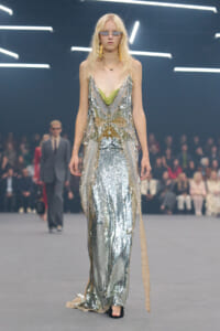 Model walking a fashion show in a silver sequined gown with gold embroidery and a lime-green neckline, wearing futuristic sunglasses.