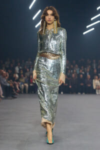 Model walking a fashion runway in a full silver sequin suit with a metallic belt and oversized gold earrings, turquoise heels visible.