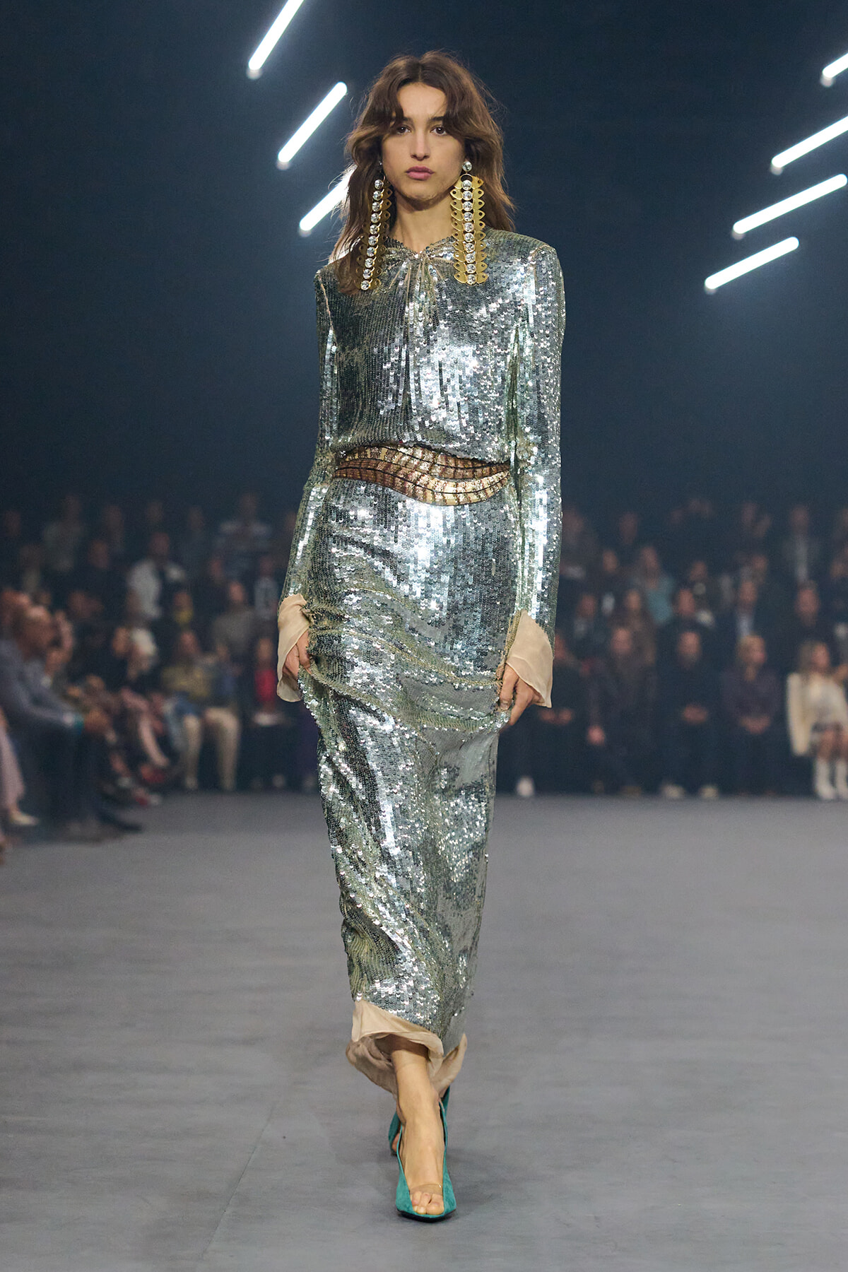 Model walking a fashion runway in a full silver sequin suit with a metallic belt and oversized gold earrings, turquoise heels visible.
