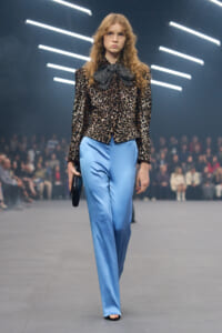 Model walking a runway in a black‑gold sequin blouse with a large polka‑dot bow, paired with light blue satin wide‑leg pants and a small black clutch.