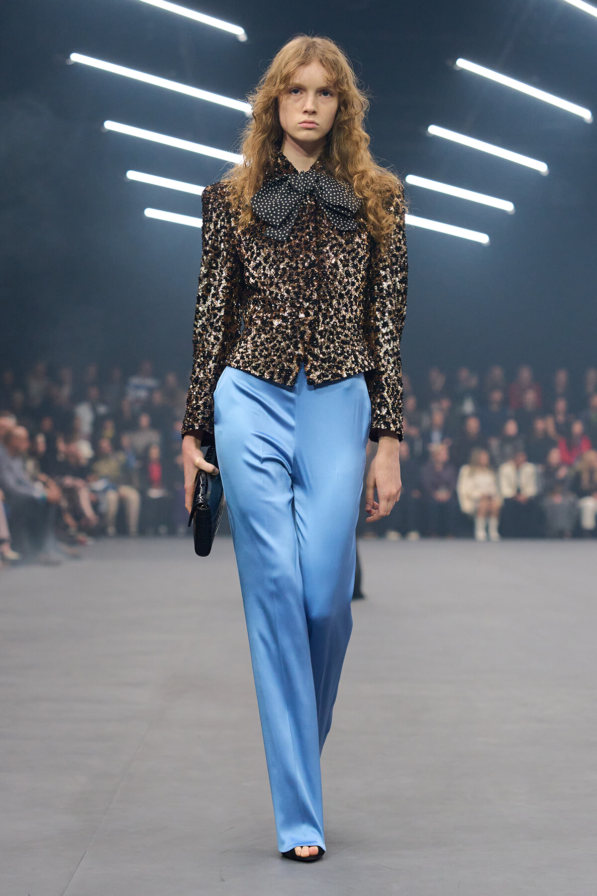 Model walking a runway in a black‑gold sequin blouse with a large polka‑dot bow, paired with light blue satin wide‑leg pants and a small black clutch.