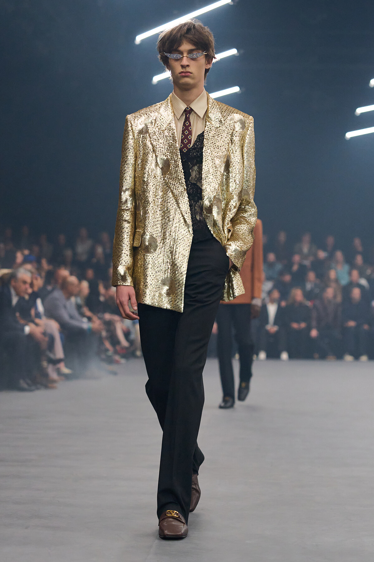 Male model on a runway wearing a gold sequined blazer, beige shirt, patterned tie, black trousers, and brown loafers with sunglasses in a dim show venue.