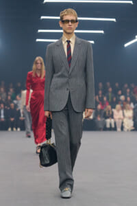 Male model in a gray pinstripe suit with a beige shirt and striped tie, wearing sunglasses, carrying a black handbag on a runway.