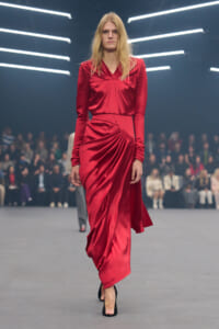 Model walks the runway in a red satin draped gown with long sleeves and black heels, audience blurred in the background.] ,
