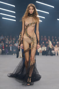 Model walks a runway in a sheer gown with a sequined snake motif, neon-lit backdrop, and audience in the background.