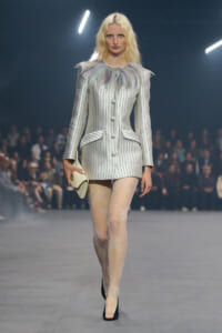 Model walks the runway in a white pinstripe blazer-dress with a feathered collar, lace tights, and a cream clutch.