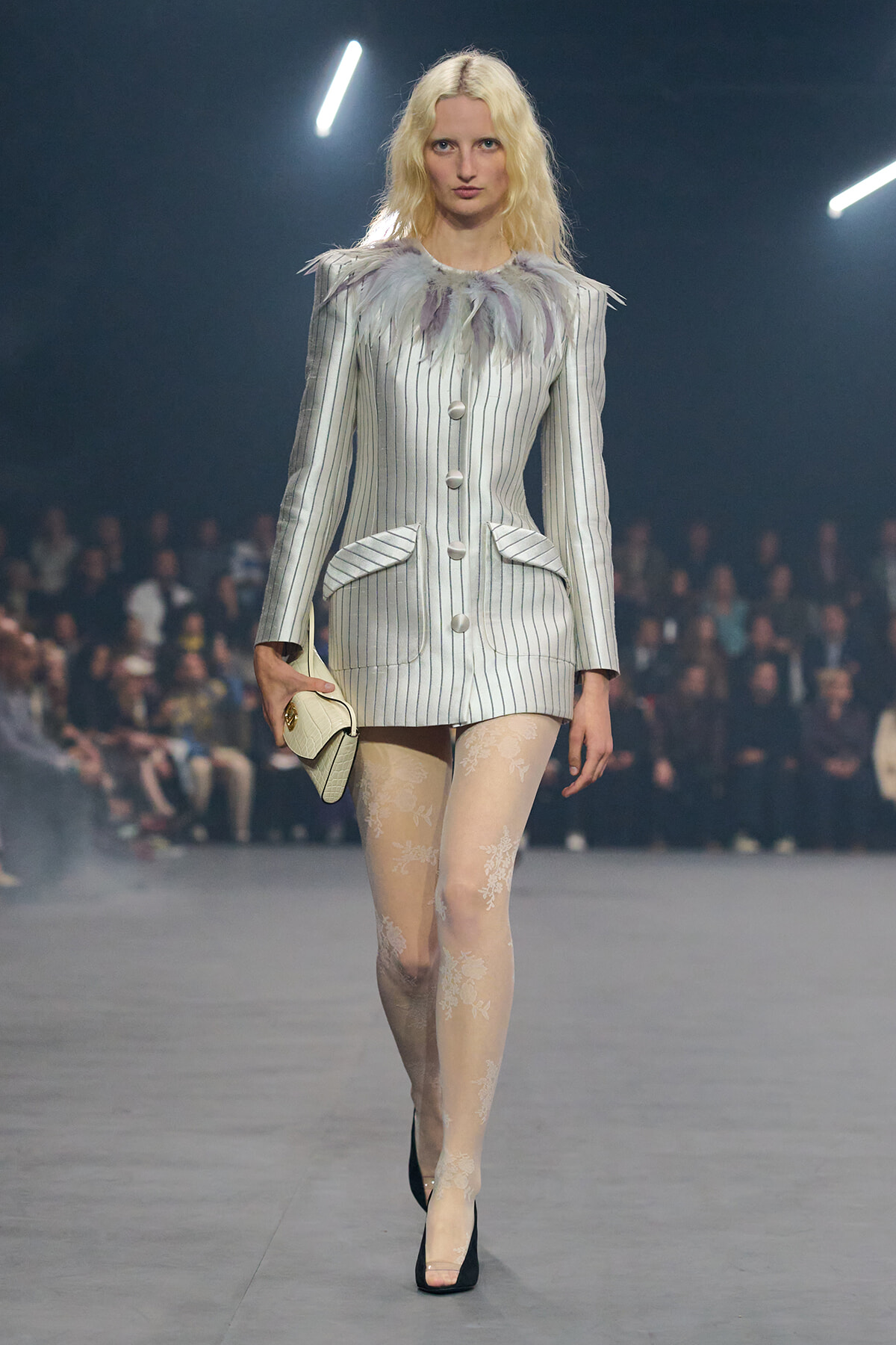 Model walks the runway in a white pinstripe blazer-dress with a feathered collar, lace tights, and a cream clutch.