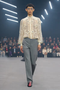 Runway look: sheer ivory embroidered blouse with floral applique, paired with gray tailored trousers.