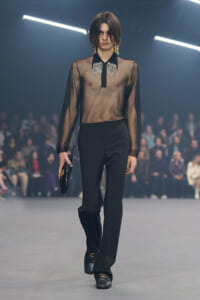 Model walking a fashion runway in a sheer black blouse with a jeweled collar, black trousers, and black loafers, carrying a small clutch.