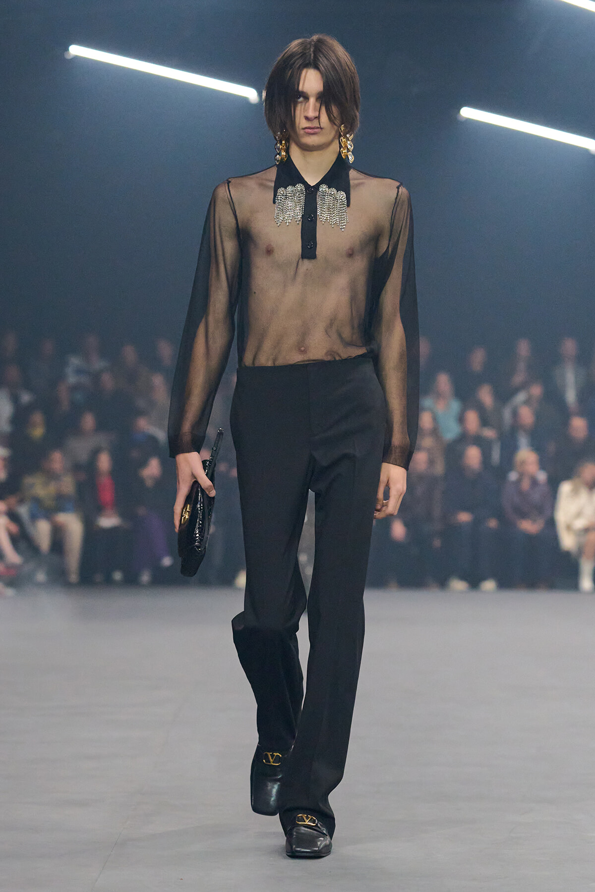 Model walking a fashion runway in a sheer black blouse with a jeweled collar, black trousers, and black loafers, carrying a small clutch.