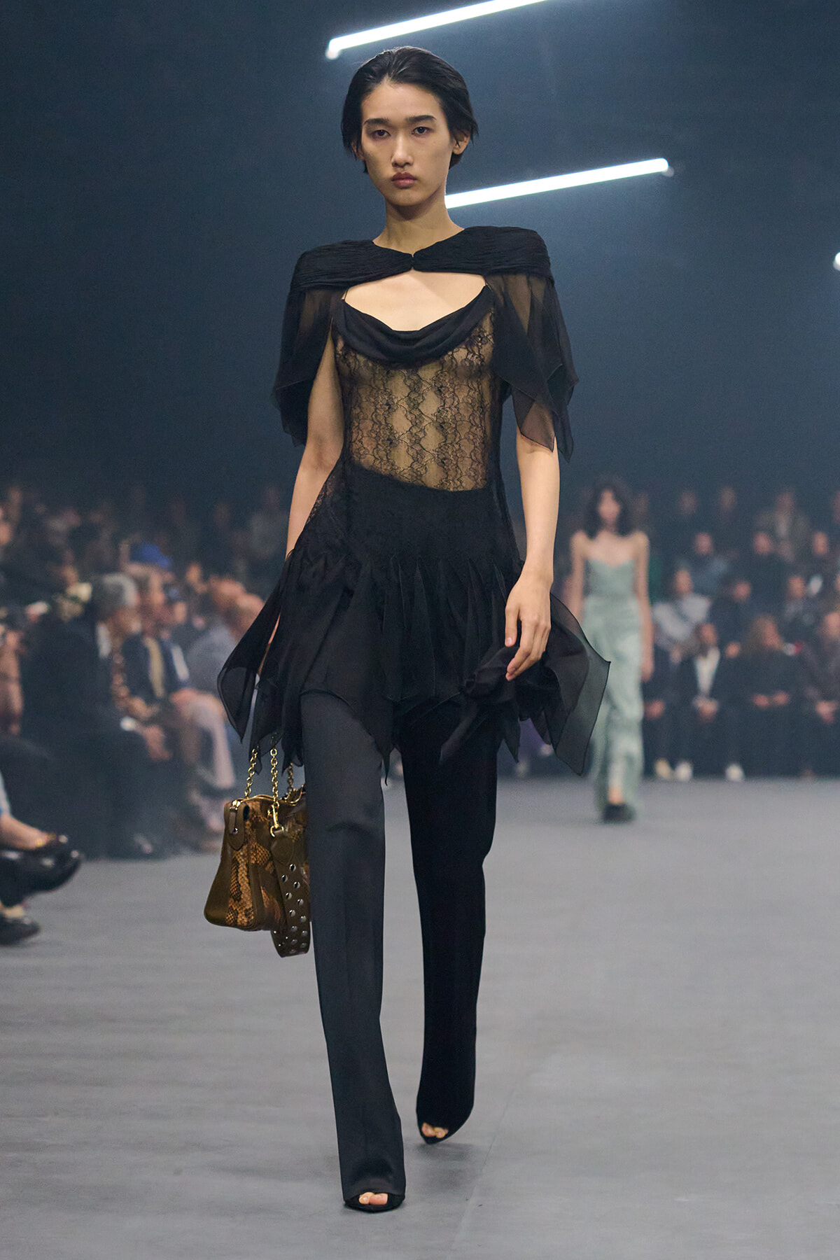 Model walking a fashion show in a sheer black lace top with cape sleeves, paired with black pants and a brown snakeskin handbag.
