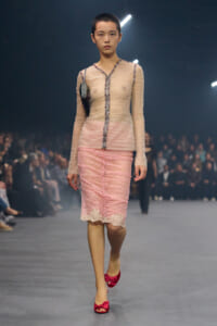 Model walks a runway in a sheer, beige-pink top with jeweled trim and a pink lace skirt; bright pink bow-heeled shoes, audience in background