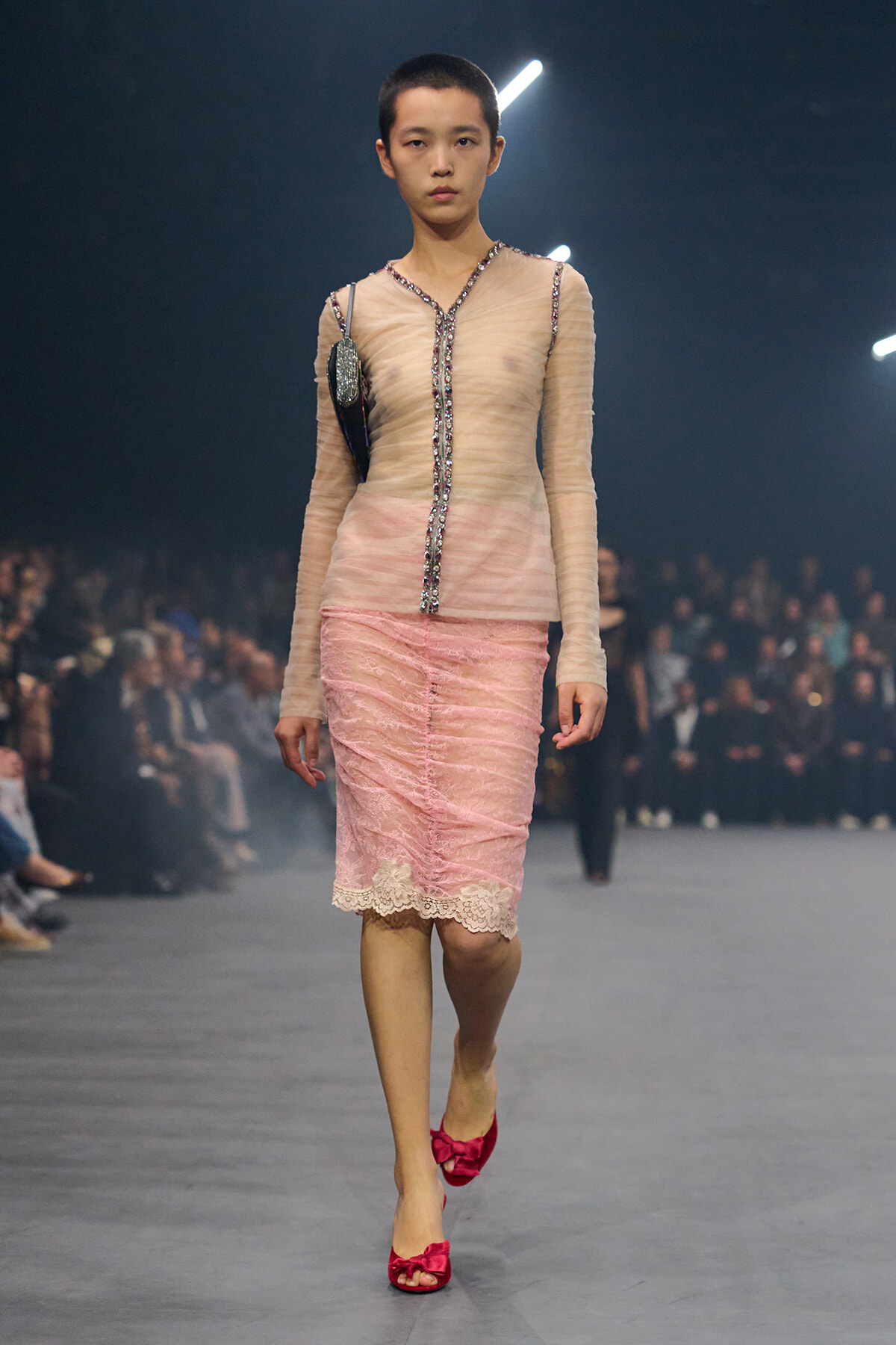 Model walks a runway in a sheer, beige-pink top with jeweled trim and a pink lace skirt; bright pink bow-heeled shoes, audience in background